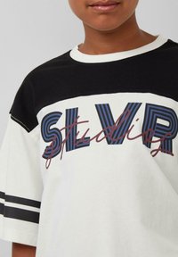 Weißes und schwarzes T-Shirt mit kurzen Ärmeln, das ein auffälliges grafisches Design in blauer und burgunderroter Schrift mit der Aufschrift "SLVR Studios" zeigt. Mit gestreiften Akzenten.