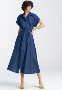 Sukienka koszulowa z denimu z krótkimi rękawami, z paskiem w talii i spódnicą w kształcie litery A do kostek. Ciemnoniebieski materiał, z bocznym rozcięciem, w połączeniu z neonowozielonymi sandalami.