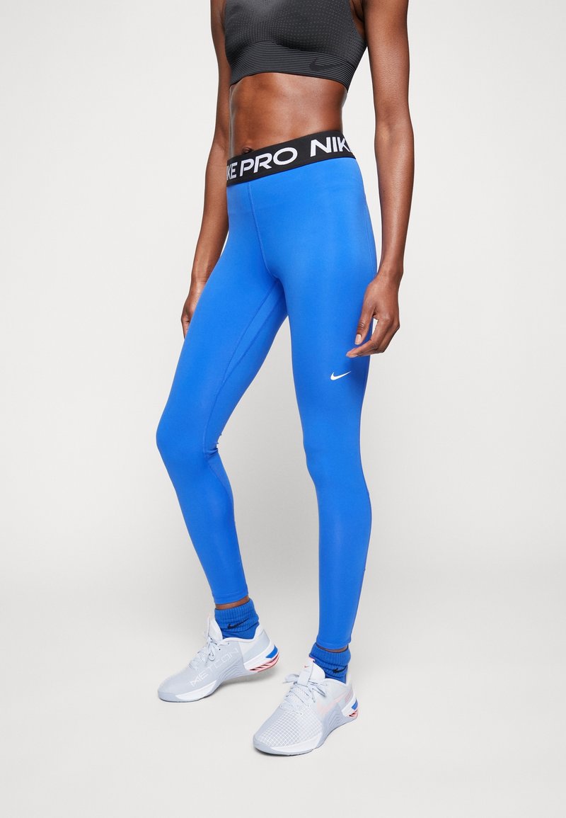 Blå Nike Pro leggings med ett svart elastiskt midjeband som har "NIKE PRO" på. Slät textur, figurnära passform, i kombination med ljusgrå träningsskor.