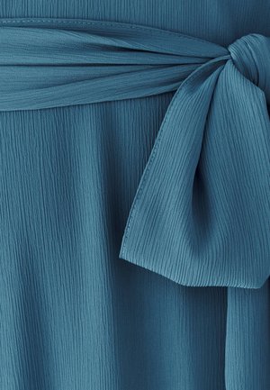 Türkisfarbener Stoff mit einer strukturierten, gerippten Oberfläche und einem gebundenen Taillendetail. Die Schleife ist locker geknotet und zeigt weiche, fließende Kanten.