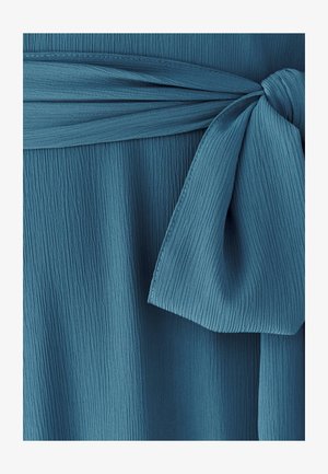 Türkisfarbener Stoff mit einer strukturierten, gerippten Oberfläche und einem gebundenen Taillendetail. Die Schleife ist locker geknotet und zeigt weiche, fließende Kanten.