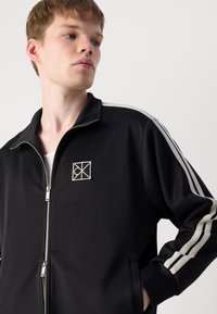 Zwarte zip-up jack met een staande kraag, voorzien van witte gestreepte accenten op de mouwen en een geborduurd logo op de borst. Gladde textuur.