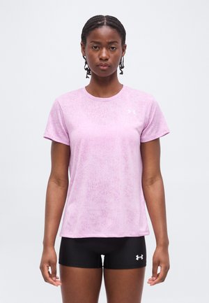 Joven mujer con el pelo trenzado, lleva una camiseta de manga corta rosa claro y pantalones cortos deportivos negros, de pie frente a un fondo blanco liso.