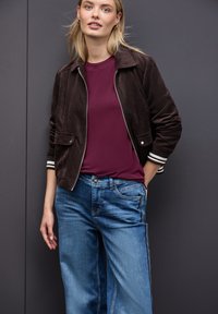 Brązowa, kordurowa kurtka na zamek z prążkowanymi mankietami, w połączeniu z ciemnofioletowym t-shirtem i wyblakłymi, szerokimi dżinsami w kolorze niebieskim.