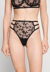 Conjunto de lingerie em renda preta apresenta padrões florais sobre uma malha transparente, com alças ajustáveis e detalhes recortados na faixa da cintura.