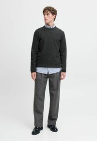 Jeune homme portant un pull gris foncé par-dessus une chemise bleu clair, un pantalon gris et des chaussures noires, se tenant devant un fond clair.