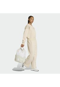 Sudadera y pantalones de chándal beige hechos de tela suave, combinados con una mochila blanca y verde claro que cuenta con correas ajustables y un bolsillo delantero.