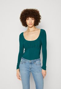 Mørkeblå langermet bodysuit med rund hals, rynkede detaljer ved brystet og tettsittende silhuett, brukt sammen med lyseblå jeans.