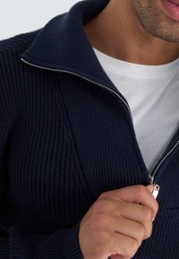 Suéter de punto azul marino con textura acanalada y cuello doblado. Presenta un zipper metálico y se lleva sobre una camisa blanca.