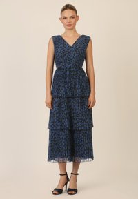 Abito blu con stampa leopardata, scollatura a V, design plisettato e strati sovrapposti. Indossato con sandali neri con tacco alto, su uno sfondo neutro.