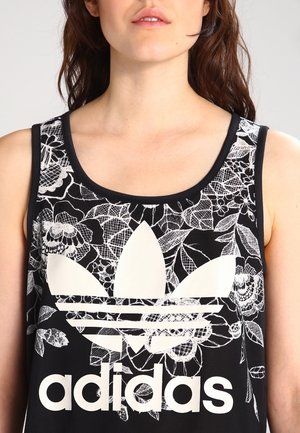 Kobieta ubrana w czarny top na ramiączkach z białymi kwiatowymi wzorami i dużym białym logo Adidas na środku klatki piersiowej.