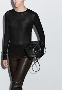 Kvinde iført sort langærmet top og mørkebrune læderbukser, der holder en sort læderhåndtaske med lynlås og rem.