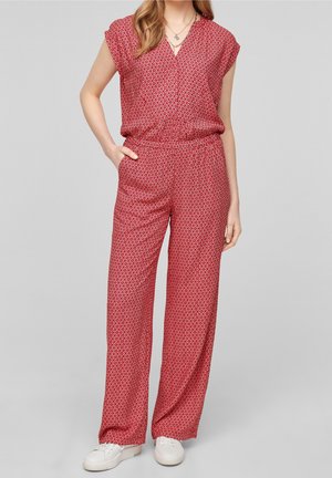 Kvinde iført en rød jumpsuit med mønster, korte ærmer og vide ben, kombineret med hvide sneakers, står med den ene hånd i lommen.
