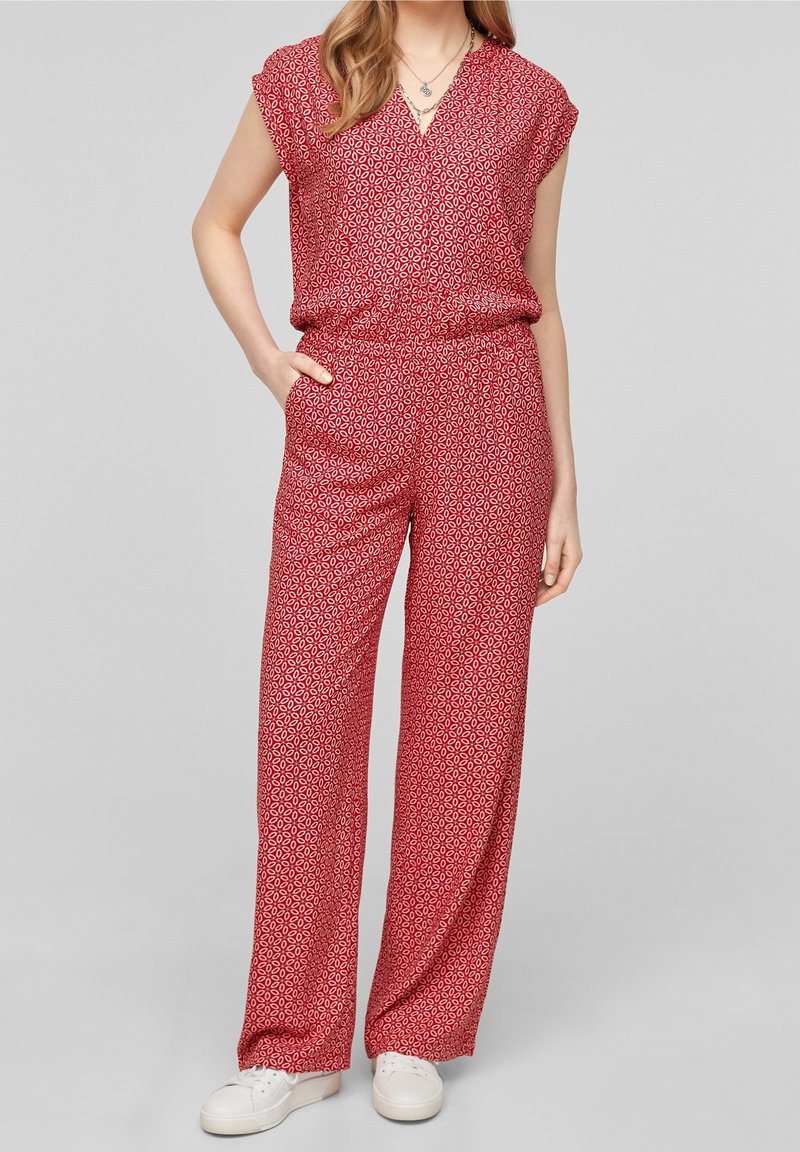 Femme portant une combinaison rouge à motifs, manches courtes et jambes larges, associée à des baskets blanches, debout avec une main dans la poche.