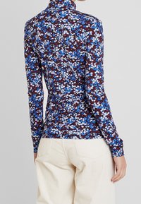 Langærmet top med høj hals, der har et blomsterprint i blå og hvid på en brun baggrund. Blødt, strækbart stof med tætsiddende design.