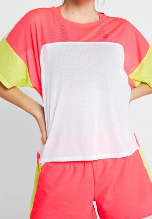 Kobieta ubrana w luźną, sportową bluzkę z krótkim rękawem w kolorach białym, koralowym i żółtym oraz pasujące krótkie spodenki w kolorach koralowym i żółtym, z rękami na biodrach.