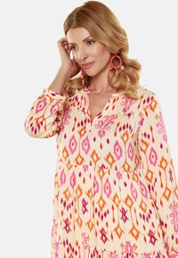 Rochie cu mâneci lungi, până la genunchi, de culoare crem, cu modele geometrice vibrante în nuanțe de roz și portocaliu, având un închizător cu butoane și un tiv în mai multe straturi.