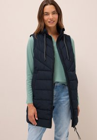 Gilet senza maniche trapuntato di colore navy con chiusura a zip e cappuccio regolabile, abbinato a una maglia a maniche lunghe verde chiaro e jeans blu.