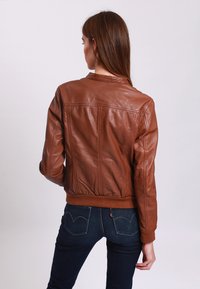 Giacca bomber in pelle marrone con polsini e fondo a costine, caratterizzata da una texture liscia e dettagli cuciti, indossata sopra jeans scuri.