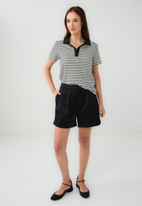 Polo a righe bianche e nere con colletto nero, abbinato a pantaloni corte neri con risvolti e ballerine nere. Outfit semplice e informale.