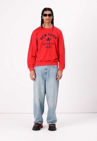 Sudadera roja con impresión negra "NEW YORK GAP ATHLETIC DEPT.", combinada con jeans de corte suelto en azul claro y zapatillas negras con detalles rojos.