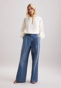Camicia bianca a maniche lunghe con scollo a chiave e bordo arricciato, abbinata a jeans in denim blu a vita alta e gamba larga. Sandali neri con tacco.