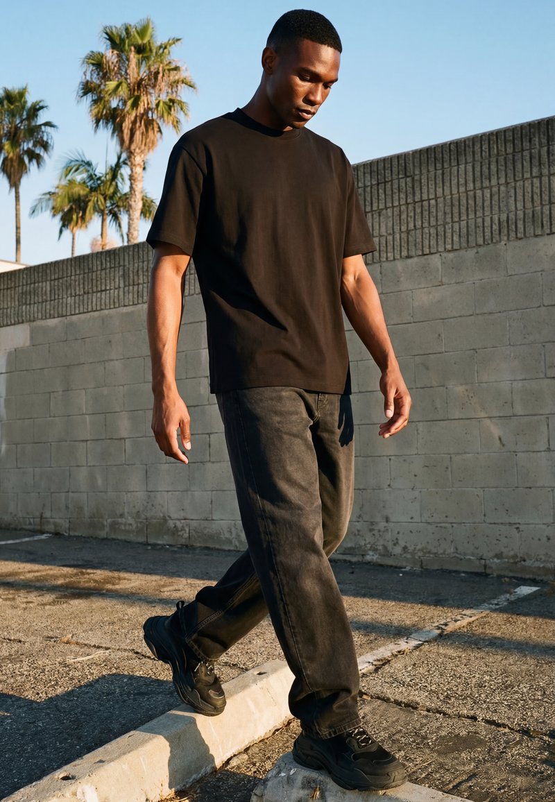 Giovane uomo con t-shirt nera, jeans e sneakers che cammina sul marciapiede accanto a un muro di cemento con palme sullo sfondo.