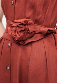 Φόρεμα λινό σε σκουριά χρώμα με ζώνη λουλούδι από ύφασμα και καφετιά κουμπιά κατά μήκος του μπροστινού μέρους, εμφανίζεται από τη μέση έως το μέσο του μηρού.