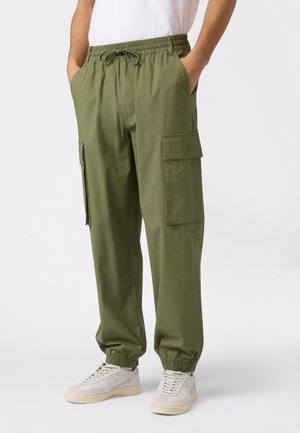 Olijfgroene cargobroek met elastische tailleband, zijzakken en elastische boorden, gemaakt van lichtgewicht stof. Gecombineerd met witte sneakers.