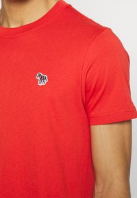 Camiseta de algodón roja con un pequeño diseño de cebra multicolor bordado en el lado izquierdo del pecho. Manga corta con un cuello redondo clásico.