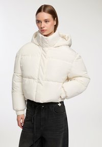 Biała puchowa kurtka z wysokim kołnierzem i kapturem, z pikowanym wzorem i elastycznymi mankietami, w połączeniu z czarnymi dżinsami.