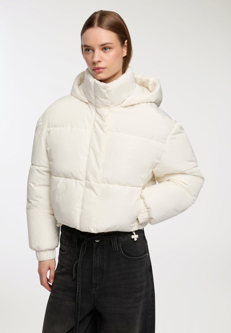 Biała puchowa kurtka z wysokim kołnierzem i kapturem, z pikowanym wzorem i elastycznymi mankietami, w połączeniu z czarnymi dżinsami.