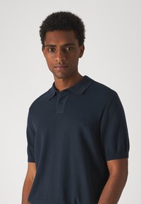 Polo bleu marine en tissu tricoté doux. Caractérisé par un col court, deux boutons et des poignets côtelés. Logo discret sur la poitrine.