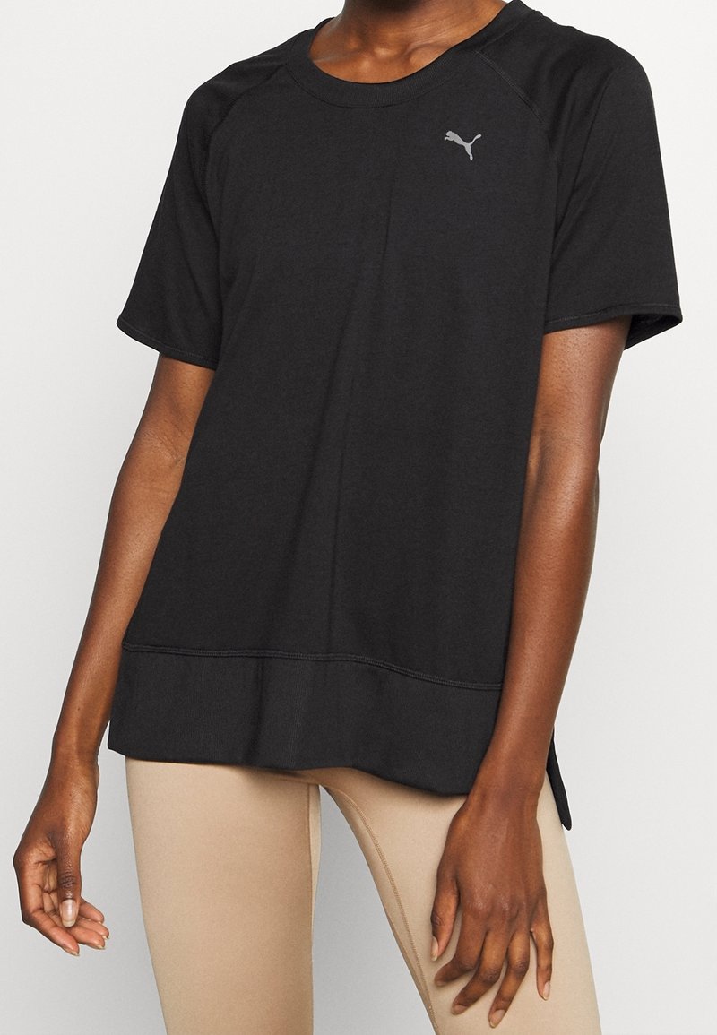 Personne portant un t-shirt noir Puma ample à manches courtes et des leggings beige, debout devant un fond clair.