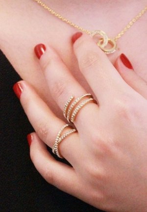 Goudkleurige ringen met verweven ontwerpen, versierd met kleine edelstenen. Circulaire hangerketting met een gestructureerde ketting. Rode nagellak.