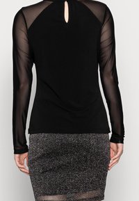 Top nero con maniche lunghe trasparenti e dettaglio a occhiello sul retro, abbinato a una gonna fitted glitterata di un grigio scuro. Tessuto liscio e vestibilità su misura.