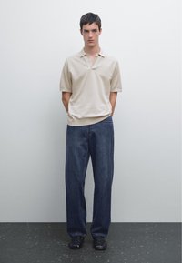 Beige pikétröja med korta ärmar, i kombination med löst sittande mörkblå denimjeans. Modellen bär svarta sandaler. Enkel bakgrund.