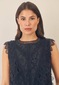 Top in pizzo blu navy con motivi floreali, rifinitura in pizzo scallopata sulle spalle e dettagli intricati lungo il collo e il corpo.
