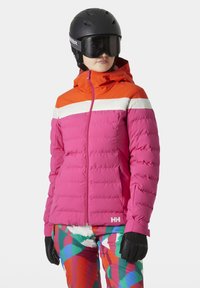 Giacca da sci imbottita rosa con cappuccio arancione e striscia bianca; presenta chiusura zip e logo, abbinata a pantaloni colorati a fantasia e guanti neri.