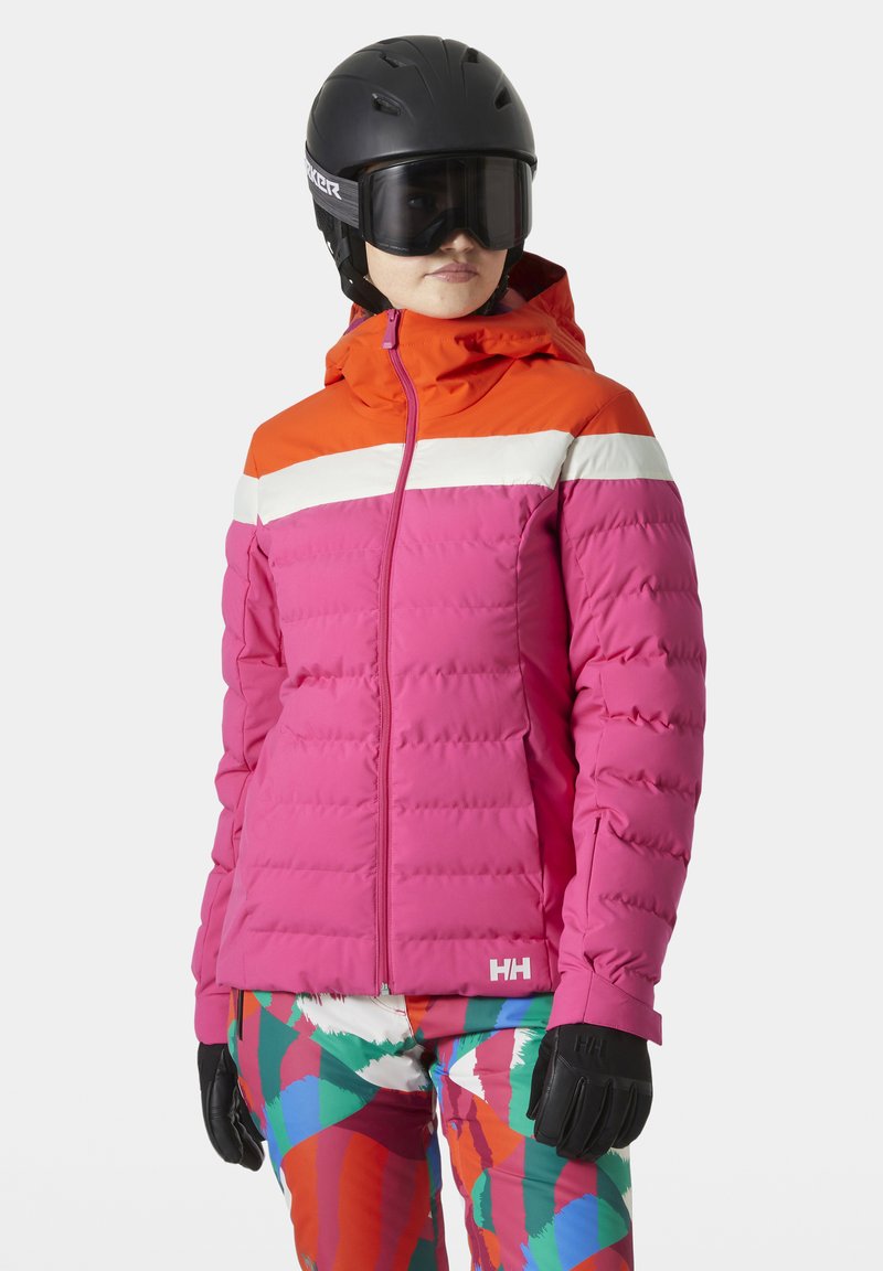 Giacca da sci imbottita rosa con cappuccio arancione e striscia bianca; presenta chiusura zip e logo, abbinata a pantaloni colorati a fantasia e guanti neri.