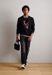 Hanorac negru cu grafică de craniu roz, blugi gri și adidași negri. Modelul ține o geantă mică neagră, mergând pe un fundal gri deschis.
