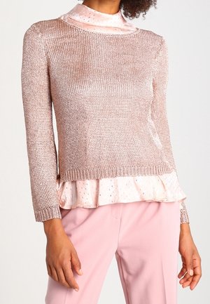 Knit sweater in metallic rose with long sleeves, layered over a pale pink ruffled blouse featuring subtle dot patterns. Worn with pink trousers.