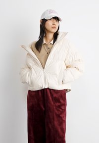 Veste crémeuse rembourrée avec fermeture éclair à l'avant, portant un polo beige et un pantalon bordeaux. La casquette blanche affiche un texte rose. Texture lisse, coupe décontractée.