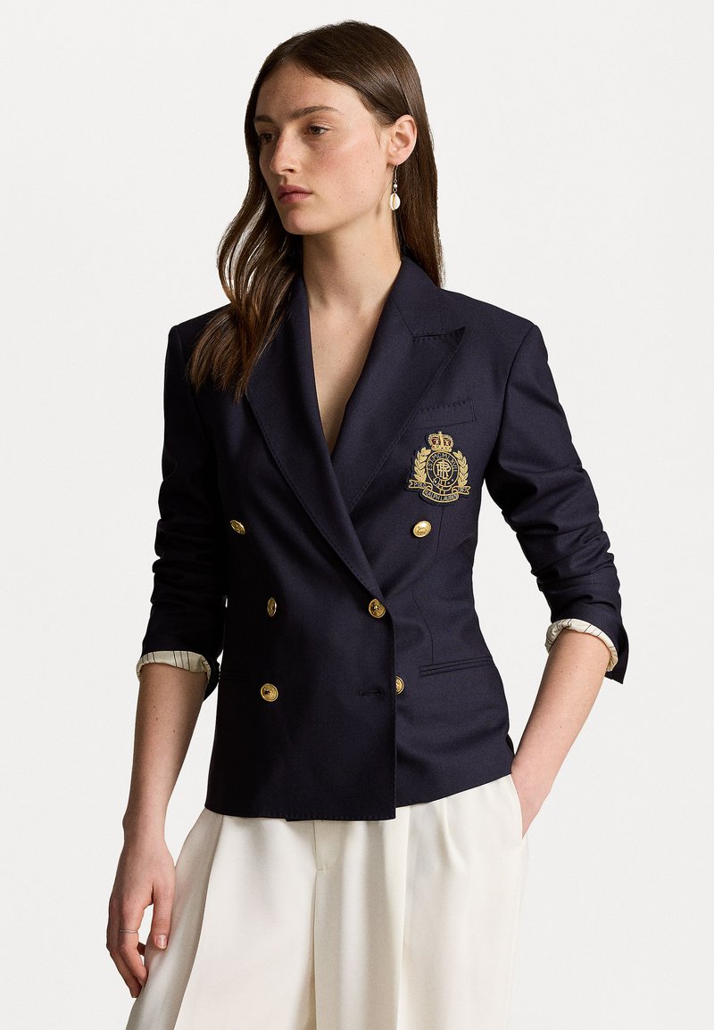 Blazer bleu marine croisé avec boutons dorés et un blason brodé. Comprend un col cranté et des manches retroussées avec une doublure à motif.