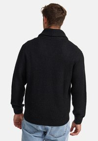 Suéter negro de punto con un acabado texturizado, que presenta un cuello de chal, puños acanalados y dobladillo, usado sobre unos jeans de color azul claro.