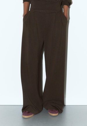 Personne portant un pantalon large marron foncé avec des plis et des poches, associé à des chaussures en daim violet avec des semelles beige.