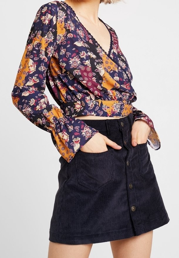 Langarm, wikkelstijl top in marineblauw en mosterdgeel bloemenpatroon, gecombineerd met een marineblauwe corduroy mini-jurk met knopen aan de voorkant en zakken.