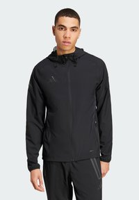 Schwarze leichte Jacke mit Reißverschluss und Kapuze, mit elastischen Bündchen und dem Adidas-Logo in reflektierender Ausführung.