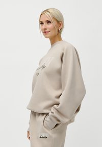Blond kvinne i beige oversized hettegenser og matchende bukser med moderne nøytralitets tekst, stående med hånden i lommen mot en hvit bakgrunn.
