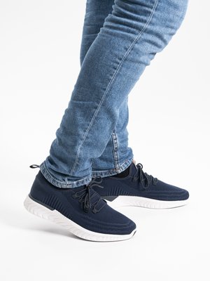 Osoba nosící tmavě modré sportovní boty a modré džíny, stojící s překříženýma nohama na bílém pozadí.